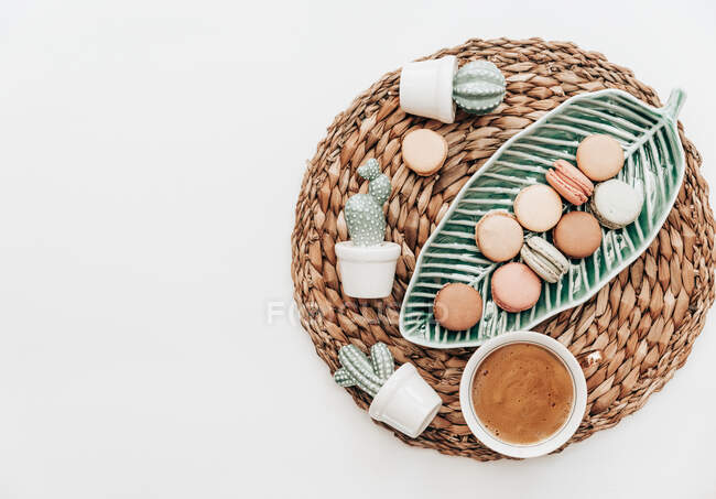 Macaroons on a leaf shaped dish, cactus decorations and a cup of coffee on a white background — Stock Photo