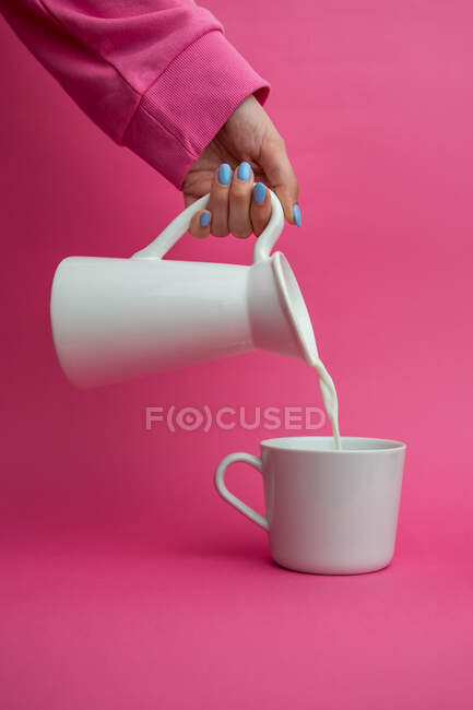 Рука протянута держа керамический кувшин и наливая молоко в чашку на розовом фоне — стоковое фото