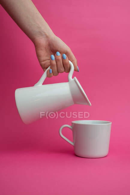 Brazo extendido sosteniendo jarra de cerámica y vertiendo leche en la taza sobre fondo rosa - foto de stock