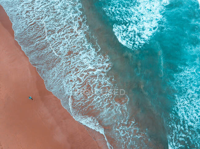Vista aérea del hombre caminando en la costa arenosa cerca del mar ondulado en el día soleado - foto de stock