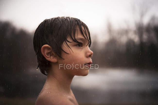 Vista lateral del niño con el pelo mojado de pie al aire libre en la lluvia - foto de stock