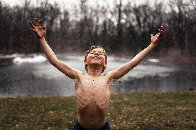 Sonriente niño de pie junto a un lago bajo la lluvia con los brazos levantados - foto de stock