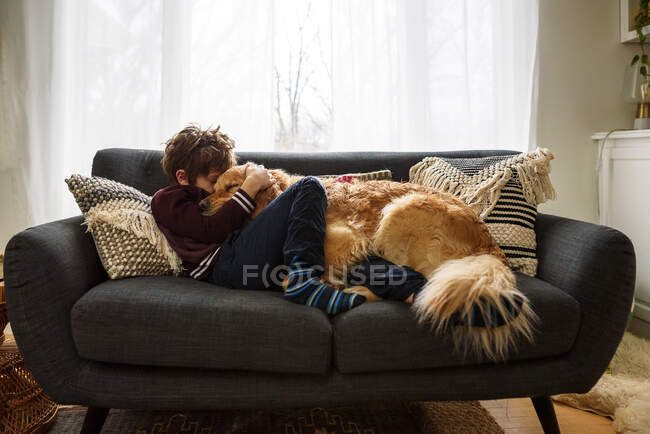 Menino deitado no sofá abraçando cão golden retriever — Fotografia de Stock