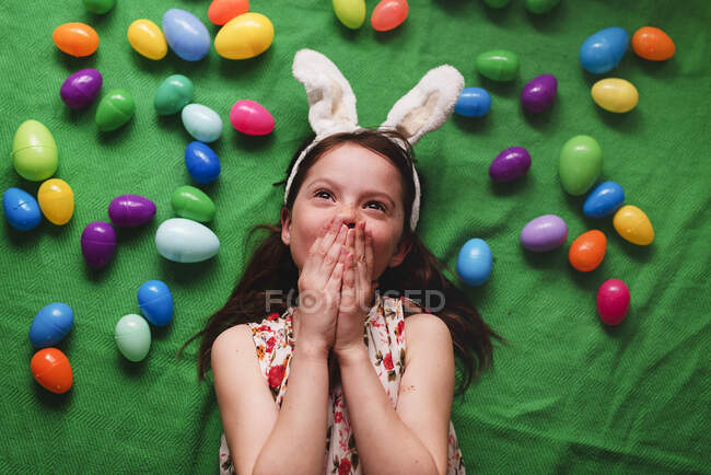 Retrato de una niña con orejas de conejo tiradas en el suelo rodeada de huevos de Pascua - foto de stock