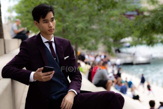 Junger Geschäftsmann sitzt am Flussufer und hält sein Mobiltelefon, Chicago, Illinois, Vereinigte Staaten — Stockfoto