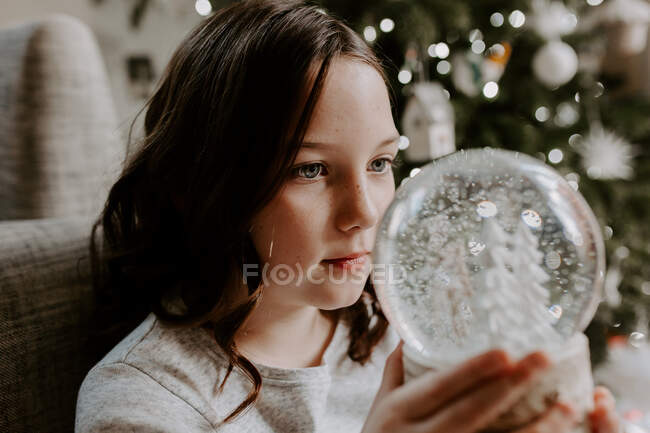 Girl sitting by a Christmas tree looking at a snow globe — Stock Photo
