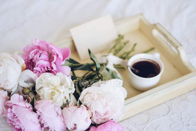 Bouquet de pivoines et une tasse de café avec une enveloppe sur un plateau sur un lit — Photo de stock