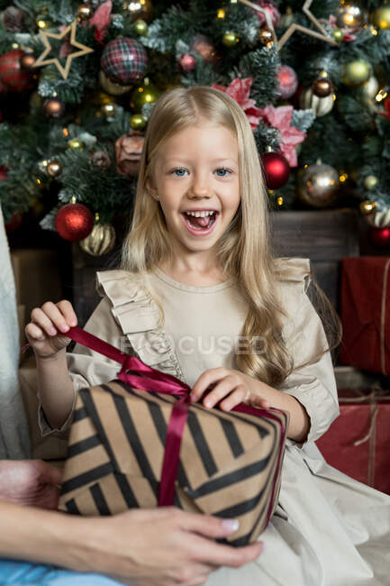 Chica sonriente sentada frente a un árbol de Navidad desenvolviendo un regalo - foto de stock