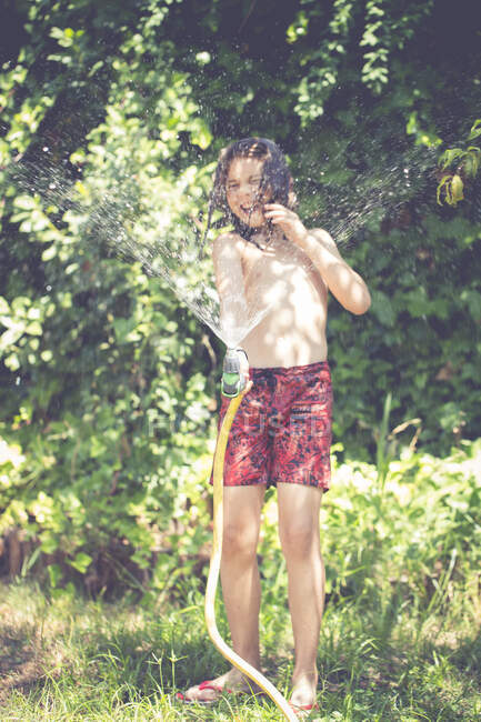 Niño feliz de pie en el jardín jugando con una manguera en el verano, España - foto de stock