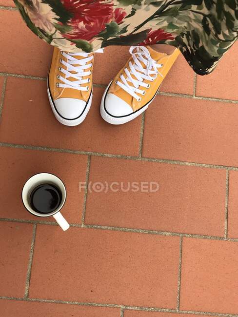 Overhead view of a woman's feet in yellow sneakers next to a cup of coffee — Stock Photo