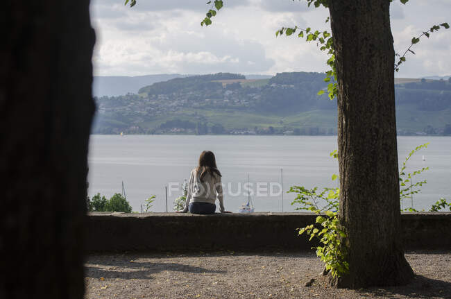 Adolescente sentada junto a un lago mirando a la vista, Suiza - foto de stock