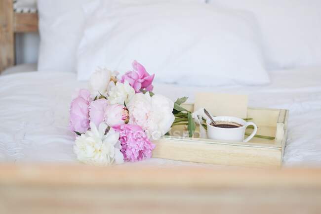 Ramo de peonías y una taza de café con un sobre en una bandeja sobre una cama - foto de stock