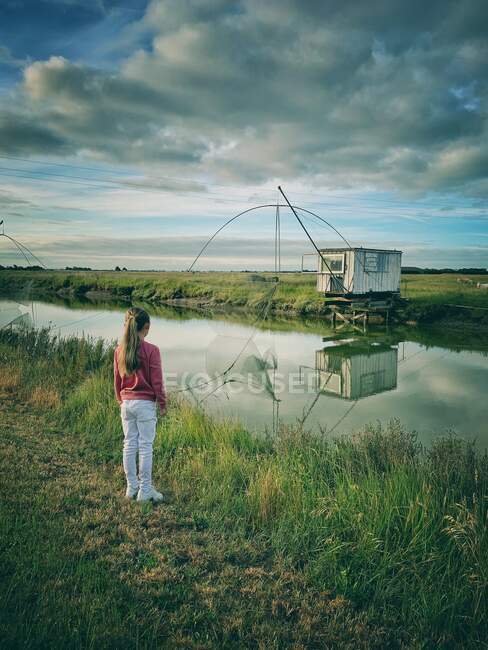 Chica de pie junto a un río mirando cabañas y redes de pesca, Rue Du Gois calzada, Isla de Noimoutier, Vendee, Francia - foto de stock