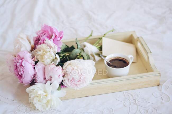 Buquê de peônias e uma xícara de café com um envelope em uma bandeja em uma cama — Fotografia de Stock