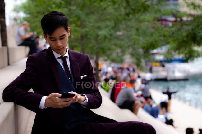 Junger Geschäftsmann sitzt am Flussufer und schaut auf sein Handy, Chicago, Illinois, Vereinigte Staaten — Stockfoto