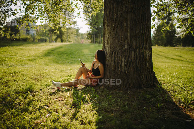 Femme assise sous un arbre regardant son téléphone portable, Serbie — Photo de stock