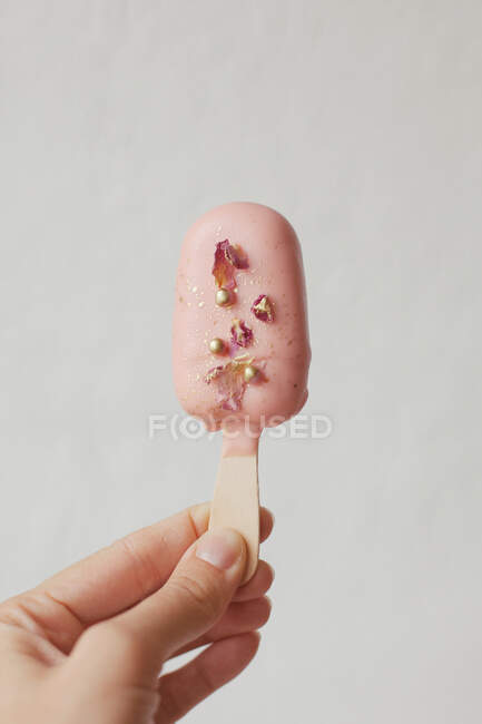 Mano de mujer sosteniendo un helado Pastel pop decorado con chispas y pétalos de rosa - foto de stock