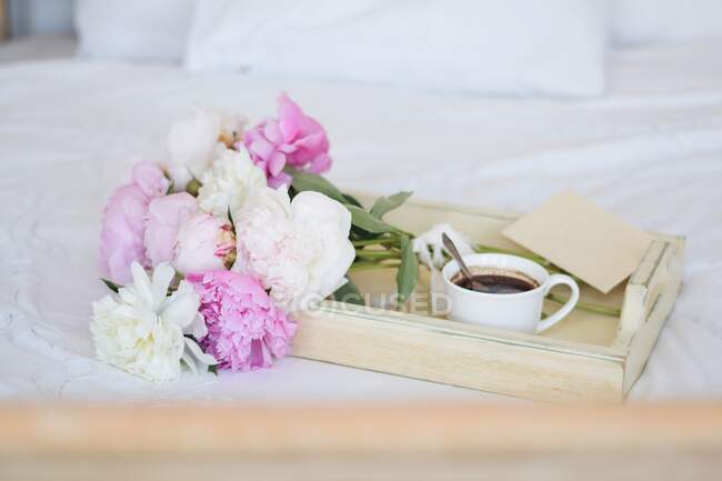 Bouquet di peonie e una tazza di caffè con una busta su un vassoio su un letto — Foto stock