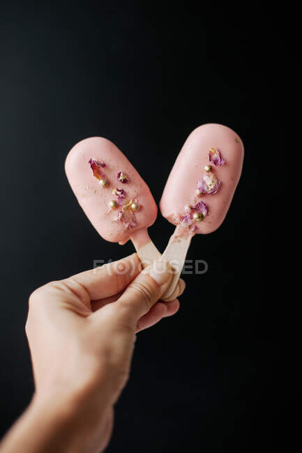 La mano de la mujer sosteniendo dos helados Cake pops decorados con aspersiones y pétalos de rosa - foto de stock