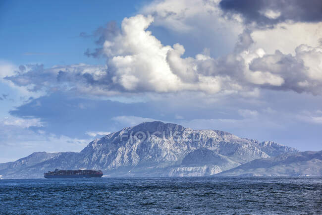Embarcación de contenedores navegando en el Estrecho de Gibraltar con la montaña Jebel Musa al fondo, Marruecos - foto de stock