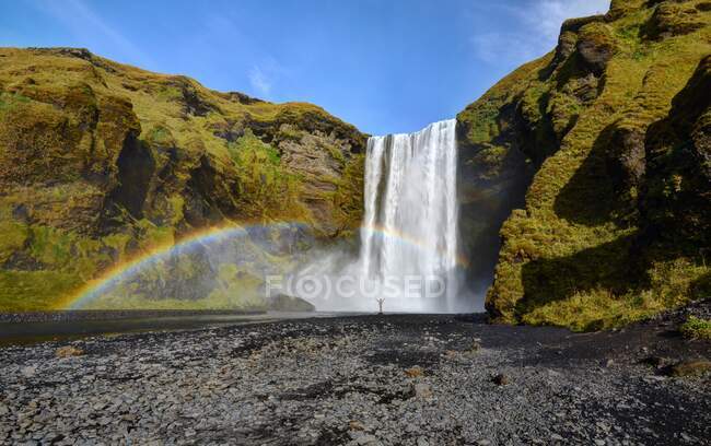 Hombre parado bajo un arco iris en la cascada de Skogafoss, Islandia - foto de stock