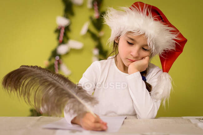 Девушка в шляпе Санты пишет письмо Санте страусиным пером — стоковое фото