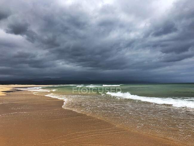 Surf waves rolling on sandy beach under cloudy sky — Stock Photo