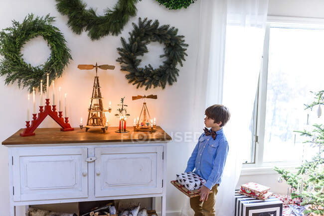Garçon debout près d'un buffet tenant des cadeaux regardant les décorations de Noël — Photo de stock