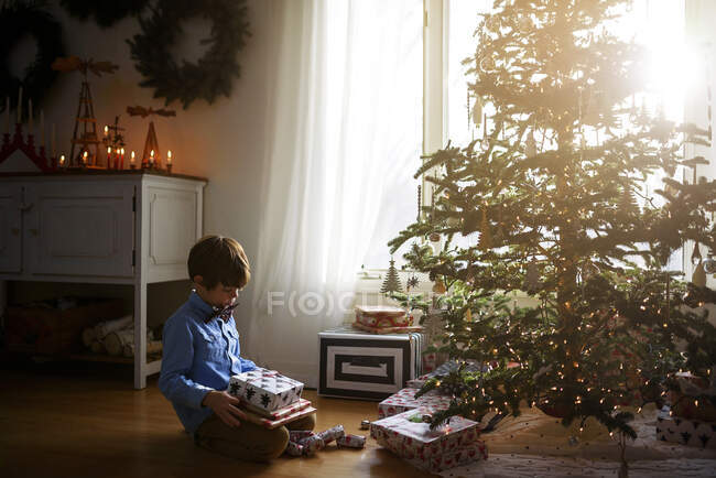 Хлопчик сидить перед ялинкою, тримаючи подарунки — стокове фото