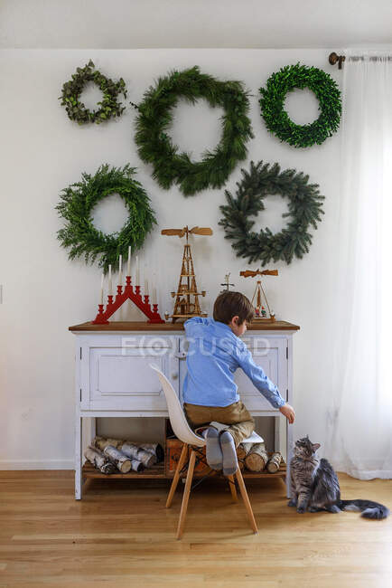 Boy kneeling on a chair in front of a sideboard stroking his cat — Stock Photo