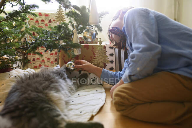 Garçon agenouillé près d'un arbre de Noël jouant avec son chat — Photo de stock