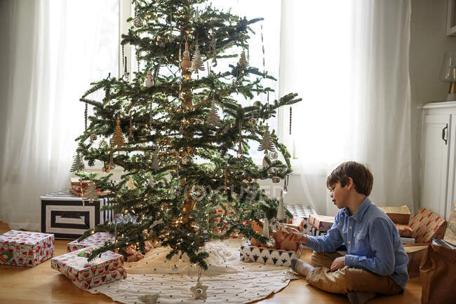 Menino sentado ao lado de uma árvore de Natal olhando para decorações — Fotografia de Stock