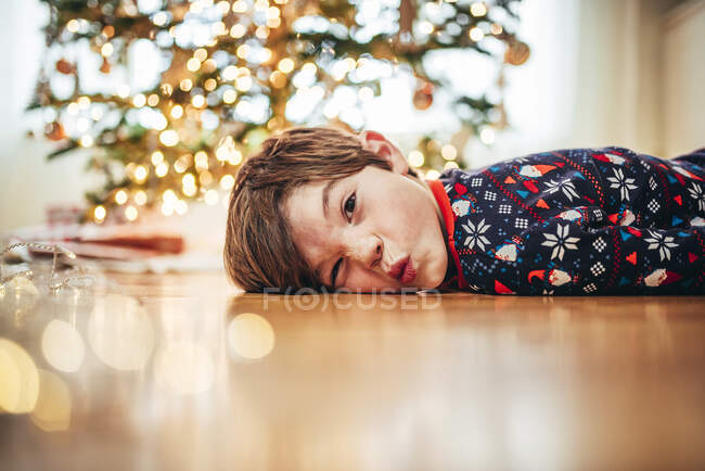 Junge liegt auf dem Boden vor Weihnachtsbaum und zieht lustige Gesichter — Stockfoto