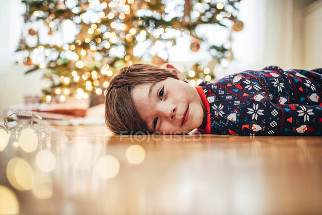 Rapaz sorridente deitado no chão em frente a uma árvore de Natal — Fotografia de Stock