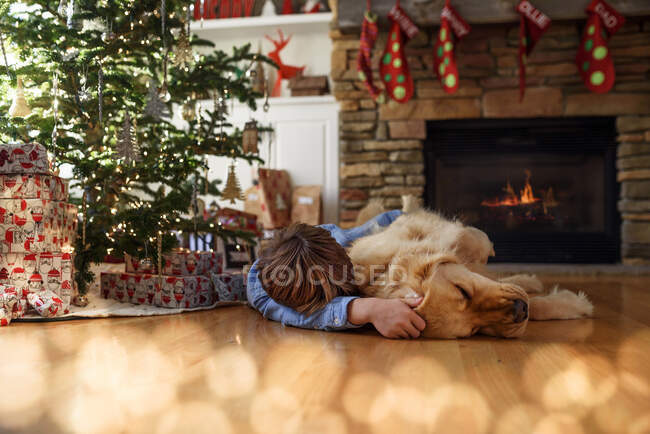 Ragazzo sdraiato sul pavimento davanti a un albero di Natale che coccola il suo cane — Foto stock