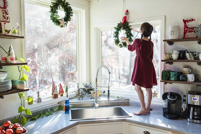 Girl standing on a kitchen worktop putting up Christmas decorations — Stock Photo