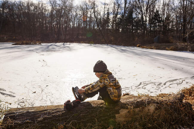 Хлопчик сидить біля замерзлого ставка і одягає ковзани. — стокове фото