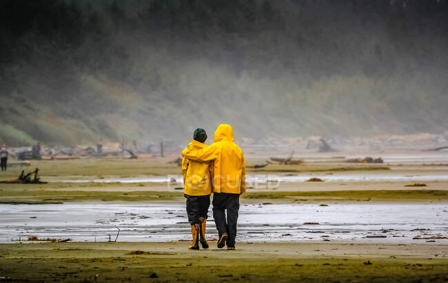 Vista trasera de una pareja caminando en una playa bajo la lluvia, Canadá - foto de stock