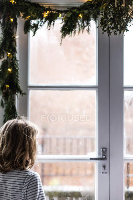 Chica mirando la nieve por una ventana decorada con abeto y luces de hadas en Navidad - foto de stock
