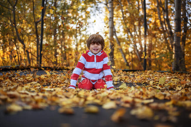 Lächelnder Junge sitzt auf einem Trampolin, das mit Herbstblättern bedeckt ist, Vereinigte Staaten — Stockfoto