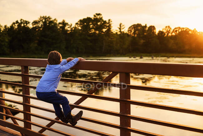 Мальчик забирается на перила моста на закате, США — стоковое фото