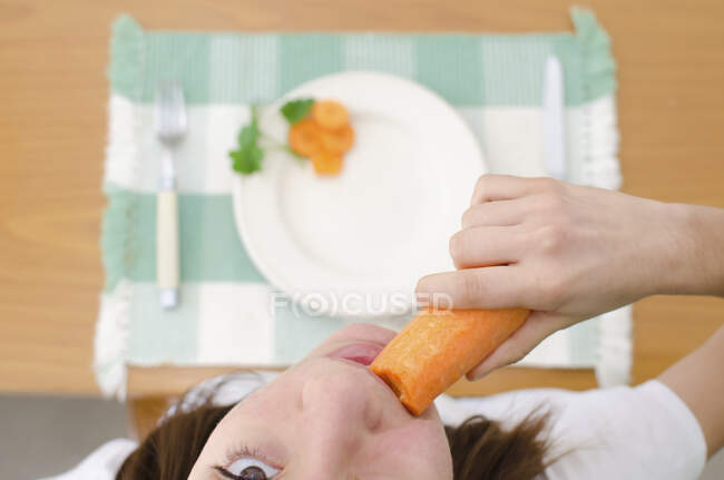 Vista aérea de una chica sentada en una mesa comiendo una zanahoria - foto de stock