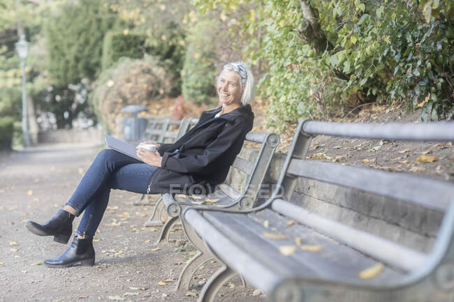 Старшая улыбающаяся женщина сидит на скамейке в парке с чашкой кофе — стоковое фото