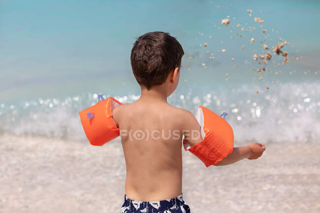 Menino em pé na praia usando braçadeiras infláveis jogando areia no ar, Grécia — Fotografia de Stock
