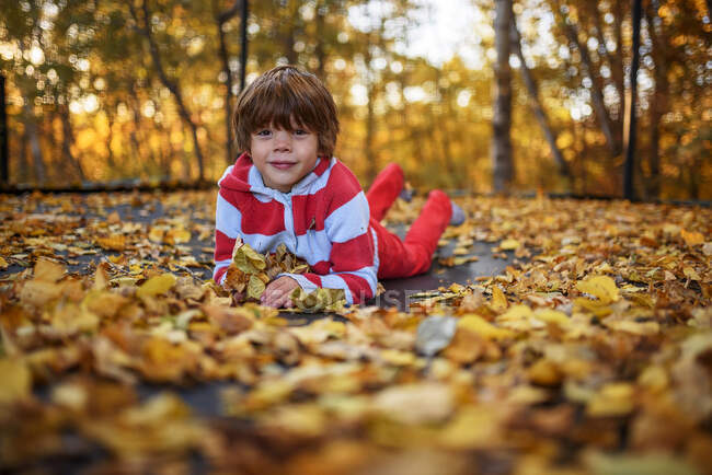 Porträt eines lächelnden Jungen, der auf einem Trampolin liegt, das mit Herbstblättern bedeckt ist, Vereinigte Staaten — Stockfoto