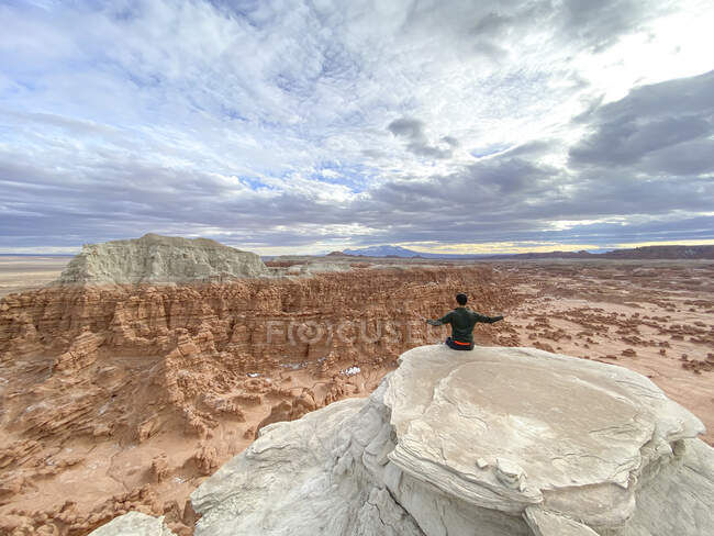 Vista trasera de un hombre sentado en el borde del acantilado con los brazos extendidos, Goblin Valley State Park, Utah, EE.UU. - foto de stock