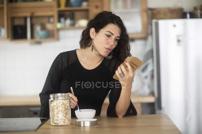 Жінка сидить на кухонній стійці, снідаючи, дивлячись на свій мобільний телефон — стокове фото