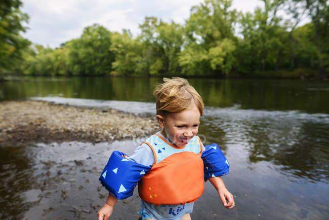Garoto sorridente vestindo um colete salva-vidas correndo ao longo de uma margem do rio, Estados Unidos — Fotografia de Stock