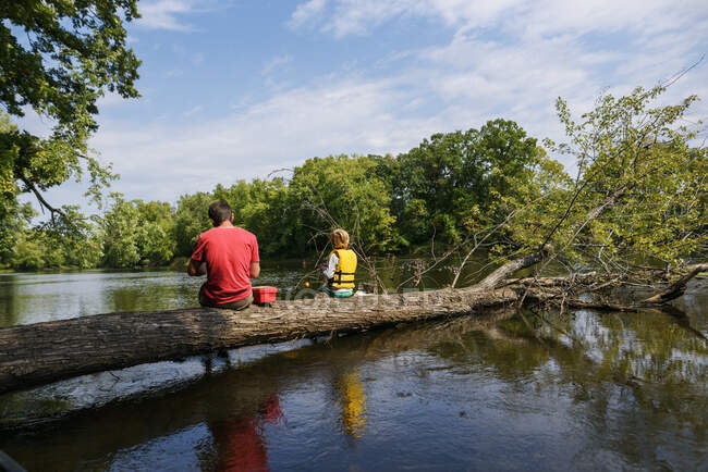 Padre e hijo sentados en un árbol caído pescando en un río, Estados Unidos - foto de stock