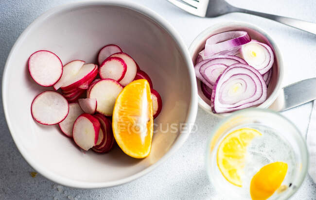Glass of lemon water and bowls of freshly sliced radishes and red onions — Stock Photo